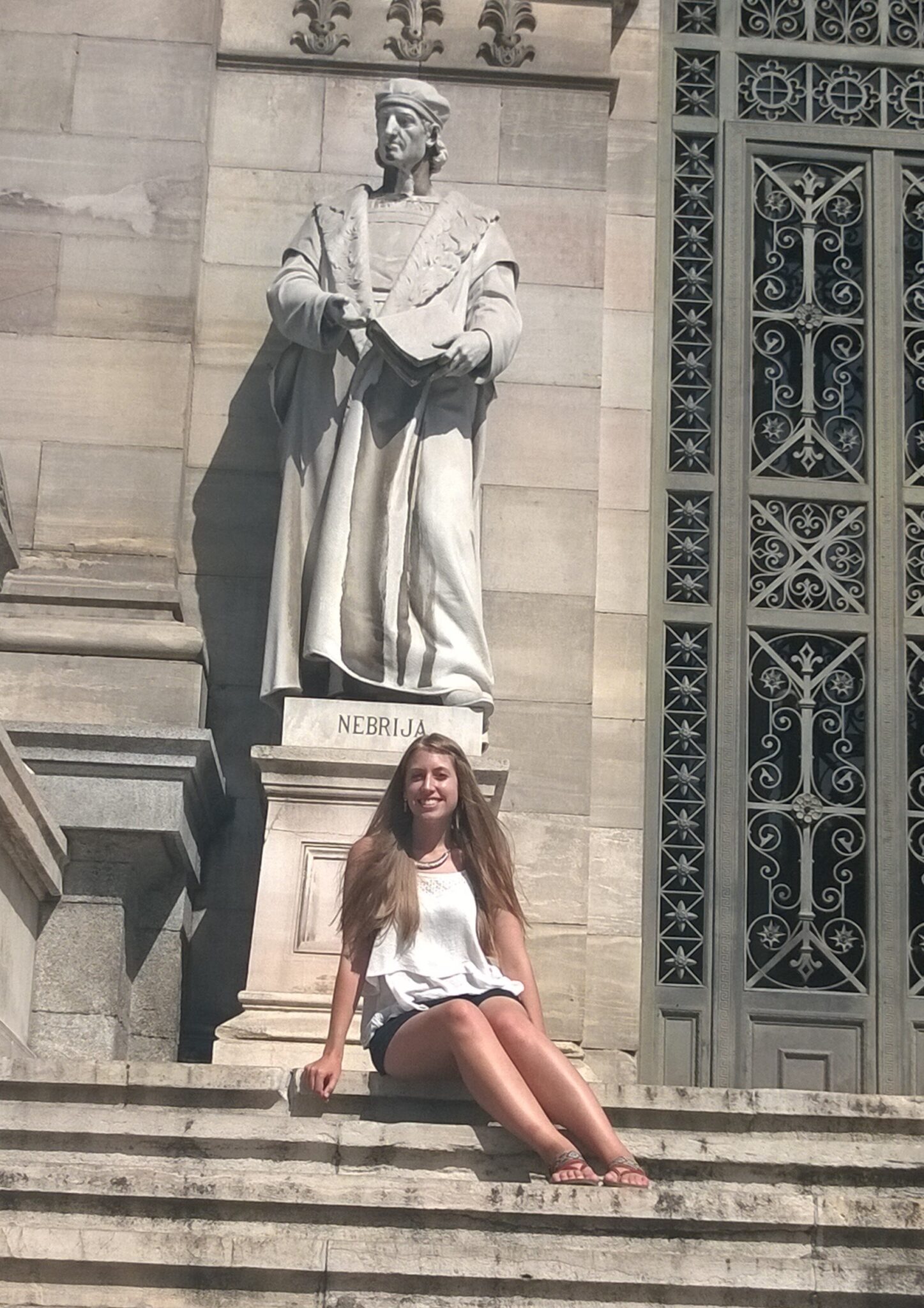 Photo shows me sitting on the steps under a large statue of a robed figure with the word "Nebrija" inscribed below.